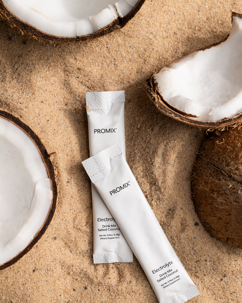 Two white Promix Salted Coconut electrolyte stick packs lying on sand between halves of a fresh coconut.