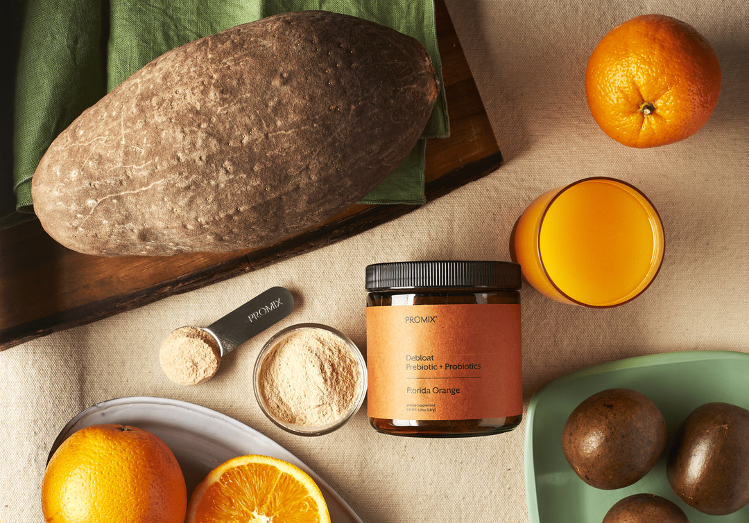 Baobab fruit beside a jar of Promix Debloat Prebiotic plus Probiotics, with milled baobab powder in a bowl, a measuring scoop, orange halves and monk fruit arranged around them.