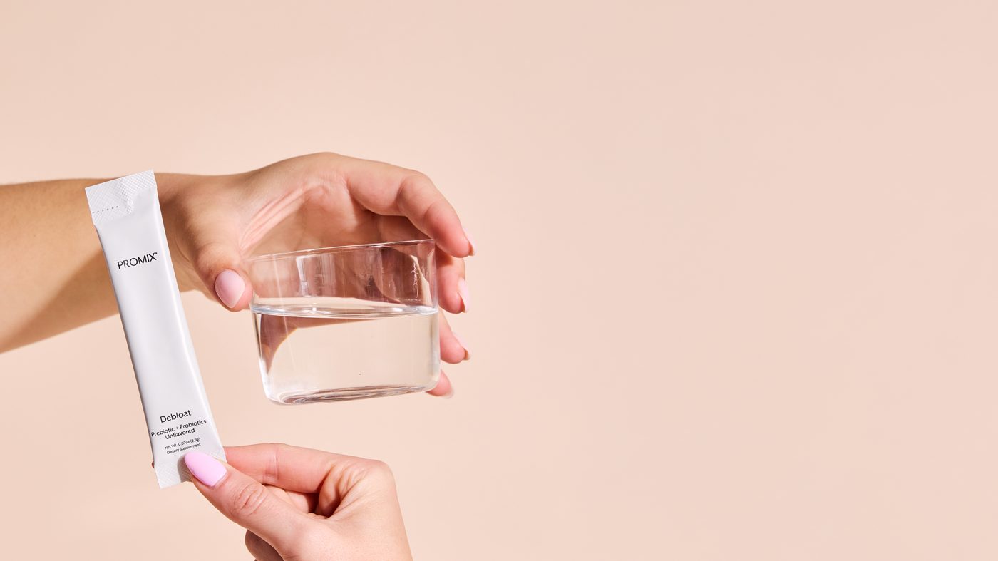 A hand holding a single Promix Debloat Prebiotic + Probiotics stick pack beside a hand holding a clear glass of water