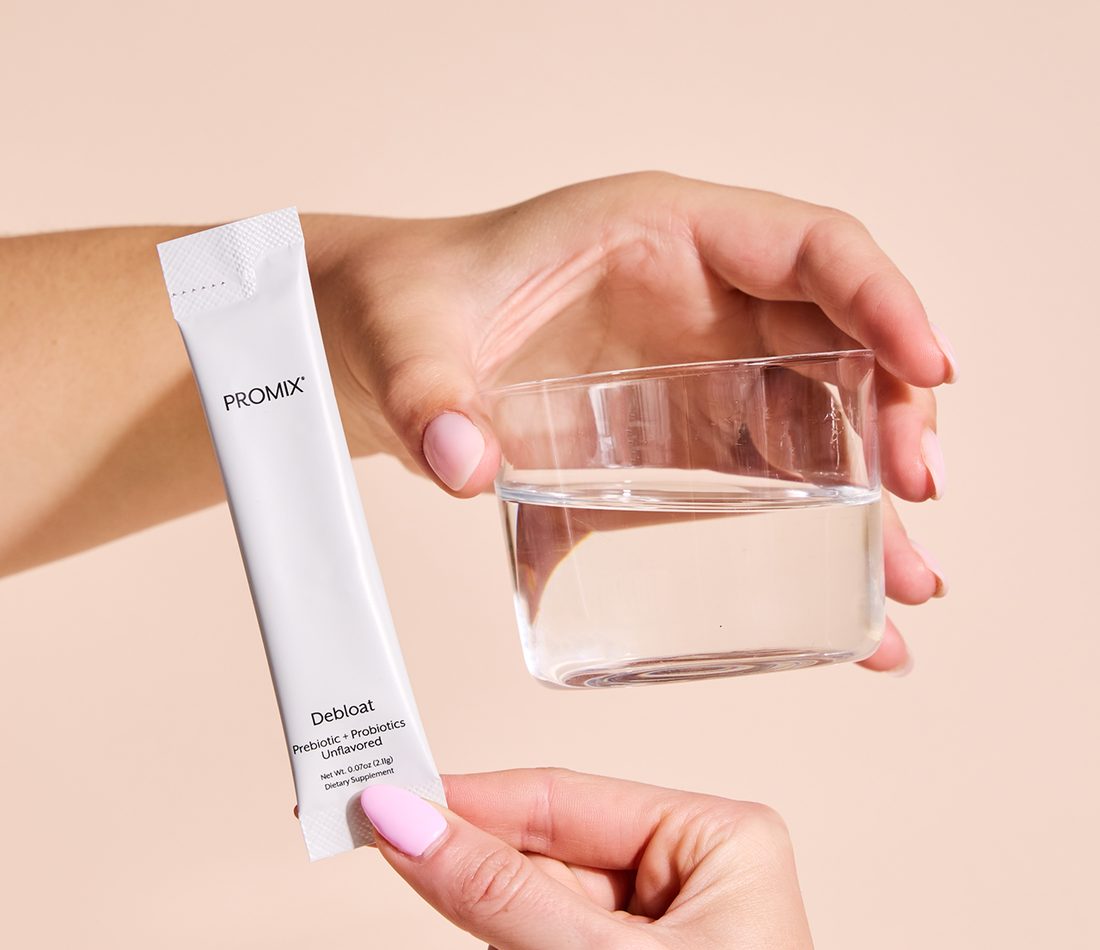 A person holding a Promix Debloat Unflavored stick pack beside a glass of water.
