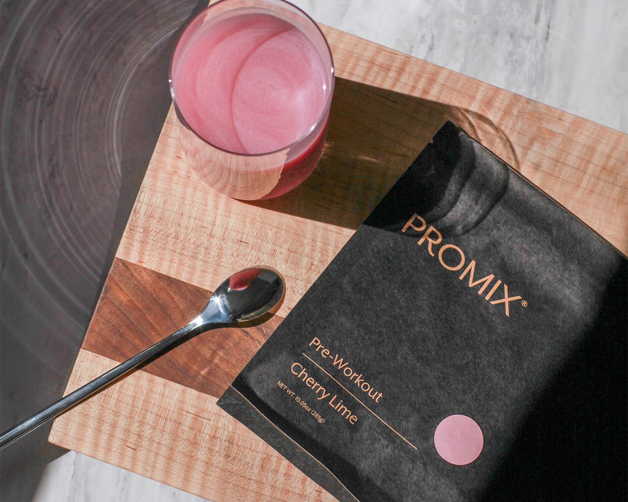 A mixed glass of Promix Cherry Lime Pre-Workout beside the pouch and a spoon on a wooden board