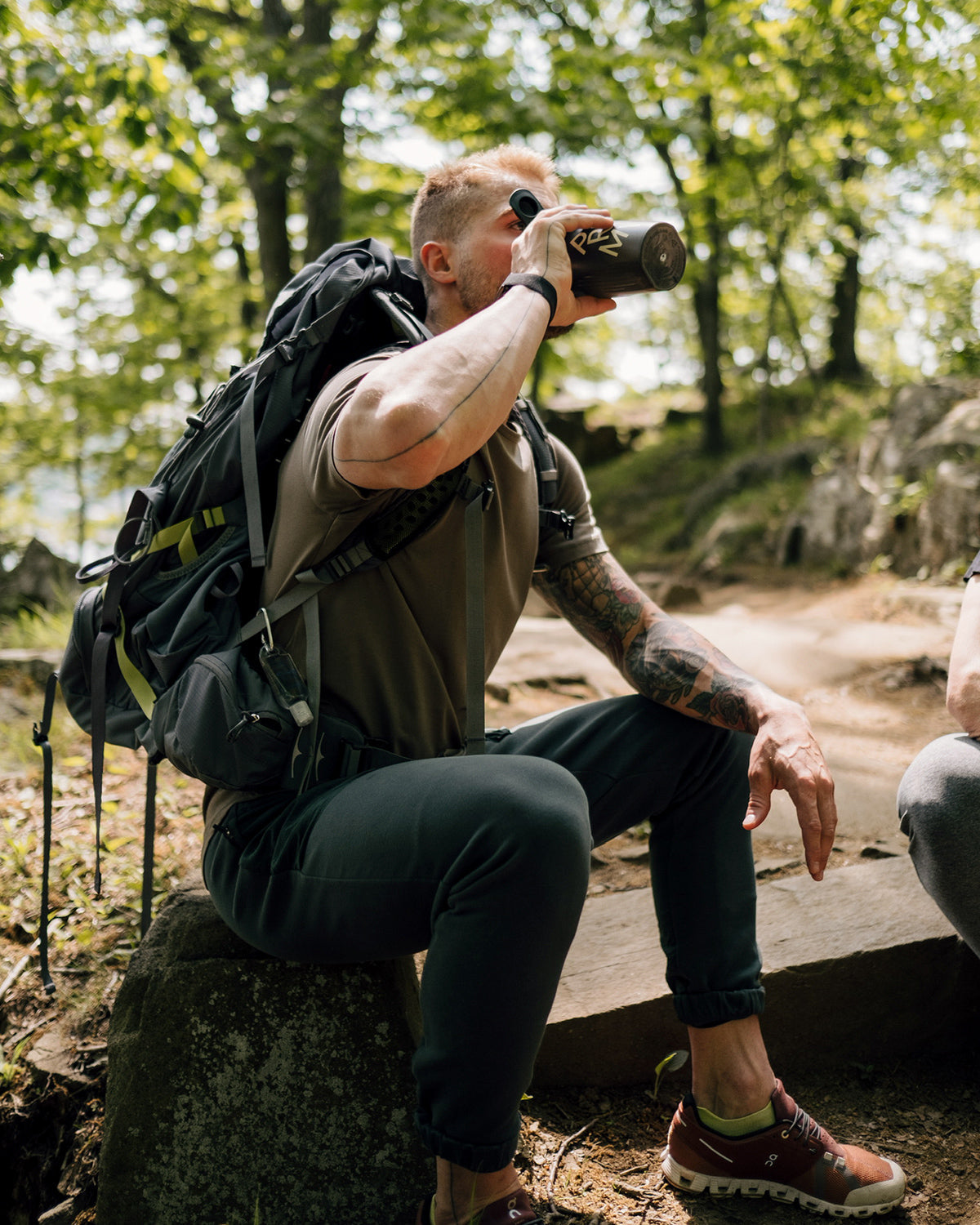 A hiker wearing a loaded backpack sits on a rock on a wooded trail, drinking from a Promix shaker bottle.