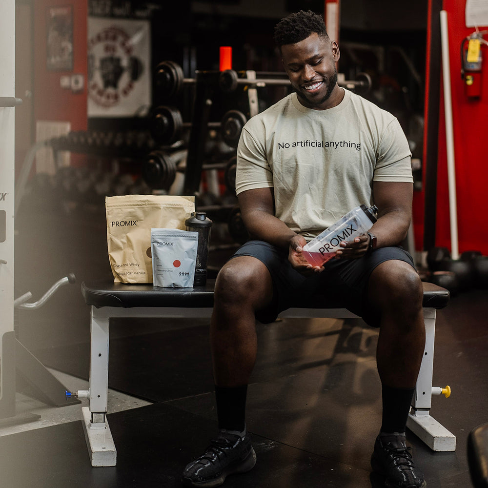 A man in a Promix No Artificial Anything shirt sits on a gym bench beside a bag of Promix grass-fed whey protein and a shaker bottle.