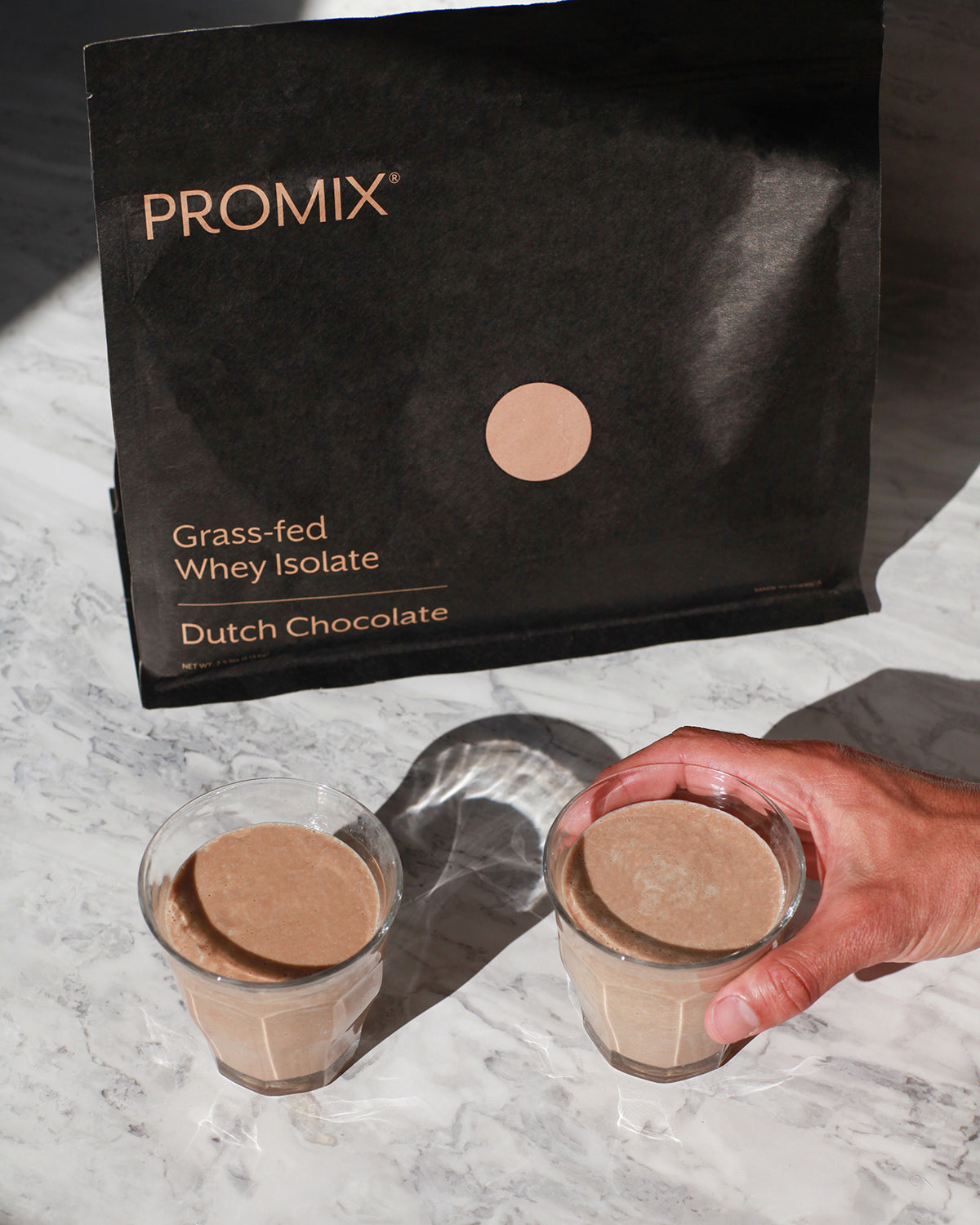 A bag of Promix grass-fed whey isolate in Dutch chocolate beside two glasses of prepared chocolate protein shake.